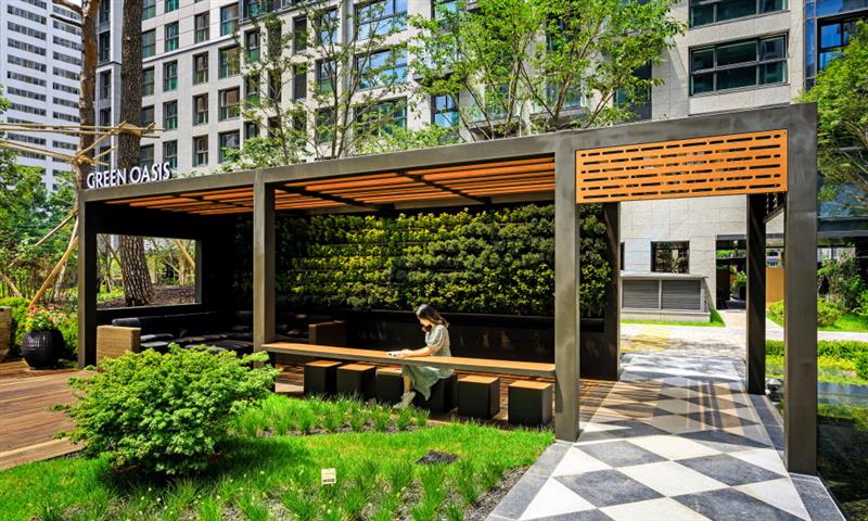 The Green Oasis Pergola, an urban relaxation space at Raemian La Classe, surrounded by modern high-rise buildings. The pergola features a wooden structure with open-air seating, lush greenery, and a paved walkway leading through the area, creating a tranquil nature escape within the city.