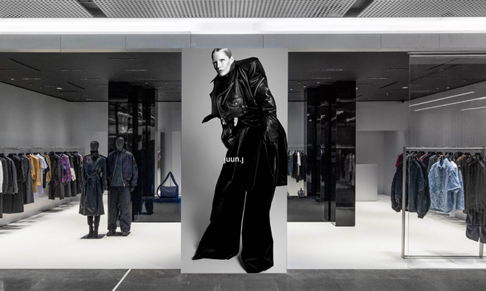 Interior of the Juun.J flagship store at Shanghai's REEL department store, featuring a monochromatic design with sleek black and white tones, clothing racks, and a striking oversized fashion visual display.