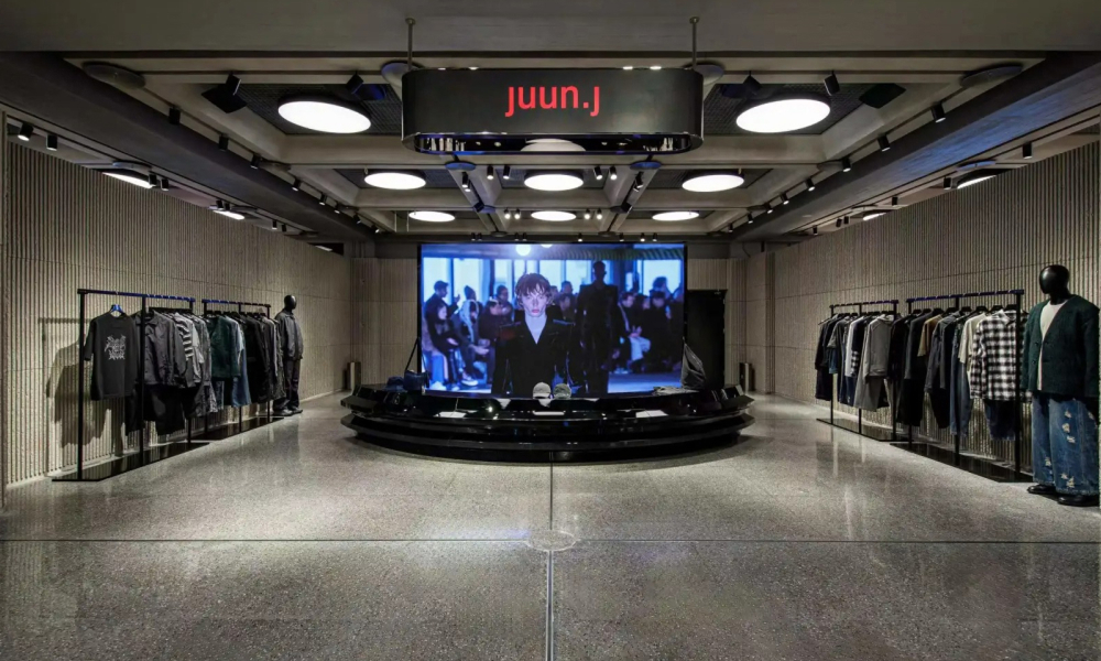 A sleek and modern Juun.J pop-up store in Chengdu, China, featuring minimalist gray interiors, clothing racks with contemporary fashion pieces, and a large screen displaying a runway video.