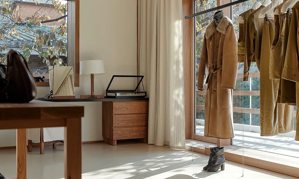 Interior of LEMAIRE's Hannam-dong flagship store featuring a minimalist design with natural wood elements, a shearling coat hanging near a sunlit window, and a desk with decorative objects in a renovated 1970s home.