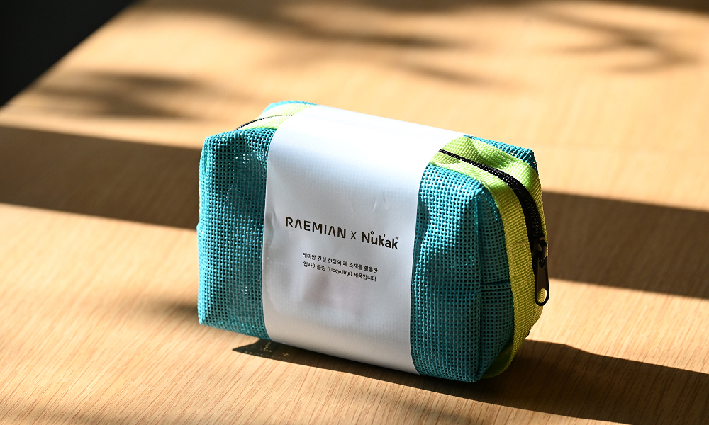 A small turquoise pouch bag with white and yellow straps, made from upcycled construction waste. The pouch, part of Samsung C&T E&C Group’s award-winning Raemian upcycled collection, sits on a wooden surface, showcasing its sustainable design and high-quality craftsmanship