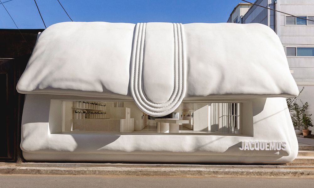 A unique Jacquemus pop-up store designed in the shape of an oversized white handbag, featuring soft, sculptural details resembling a luxury purse. The storefront has an open entrance, allowing a glimpse of the minimalist interior. The setting is in an urban environment with visible power lines and buildings in the background.