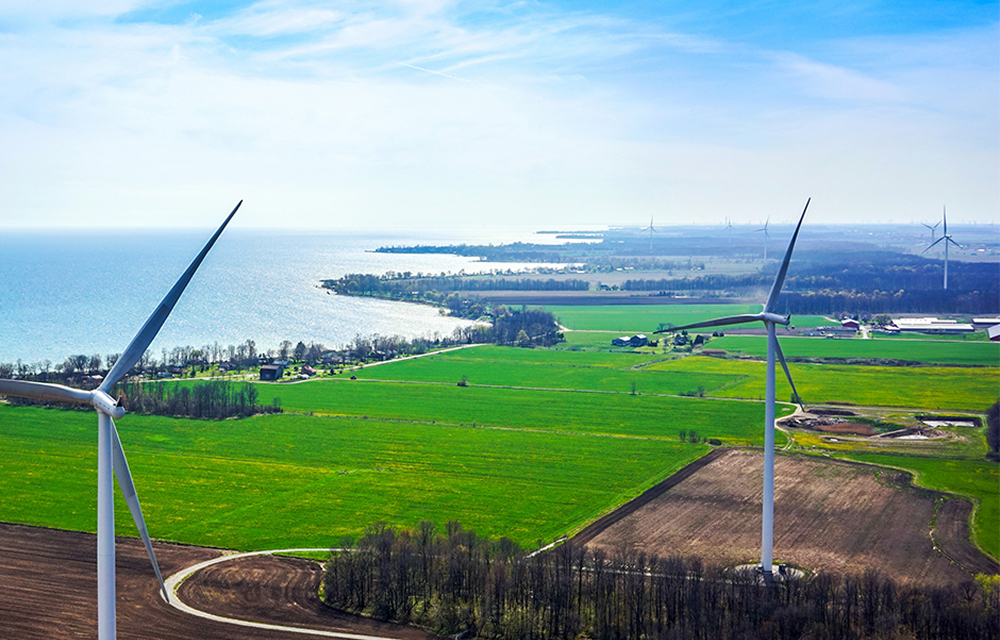 Aerial view of the North Kent Wind Farm in Ontario, Canada, showing wind turbines scattered across green agricultural fields near a lake on a bright, clear day.