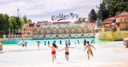 Visitors run into the large outdoor wave pool at Caribbean Bay under a bright spring sky, with the park’s signature sign and Mediterranean-style backdrop visible in the distance.