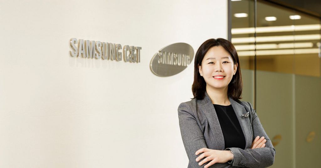 Jungwan Kim poses with arms crossed, smiling beside a wall with the Samsung C&T logo at company headquarters.