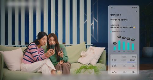 Two women sitting on a sofa, looking at a smartphone together, with an on-screen energy usage comparison graph displayed from Samsung C&T E&C Group’s smart home platform.