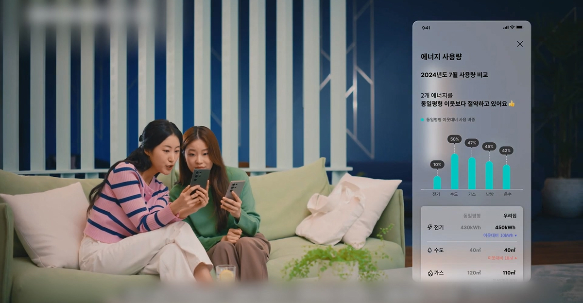 Two women sitting on a sofa, looking at a smartphone together, with an on-screen energy usage comparison graph displayed from Samsung C&T E&C Group’s smart home platform.