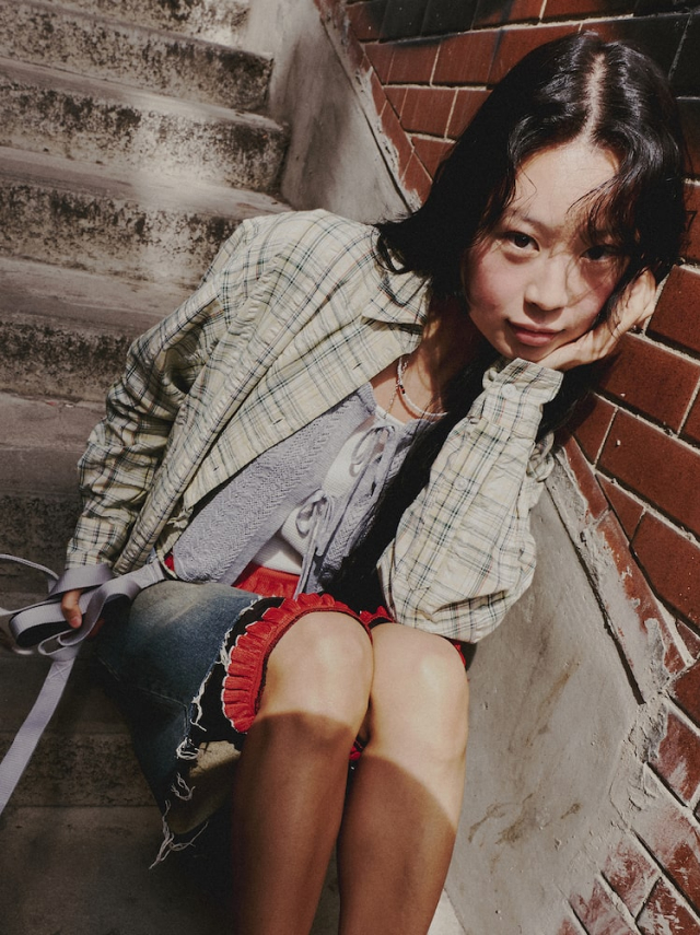 Jisoo sits casually on the stairs styled in a plaid shirt, layered knits, and knee-length jorts.
