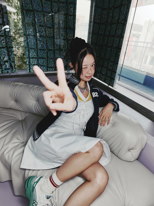 Jisoo Seo lounges on a couch holding up a peace sign wearing a white dress layered under a navy zip-up jacket, accessorized with striped socks, and green sneakers.