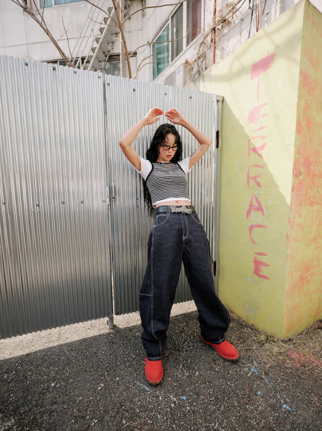 Jisoo poses outdoors in front of a metal fence, wearing a striped tank top over a white tee and wide leg jeans, paired with bold red shoes.