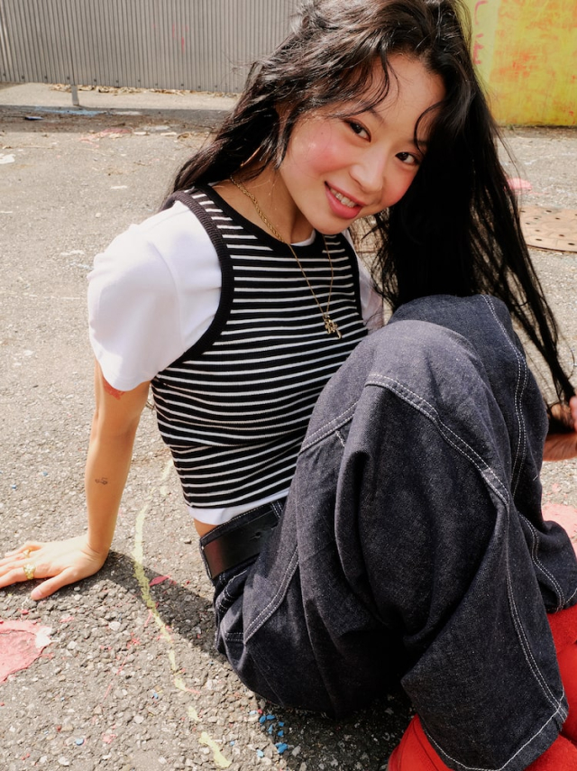 A close-up of Jisoo Seo sitting on the pavement in a striped tank layered over a white tee, dark denim pants, and red shoes.