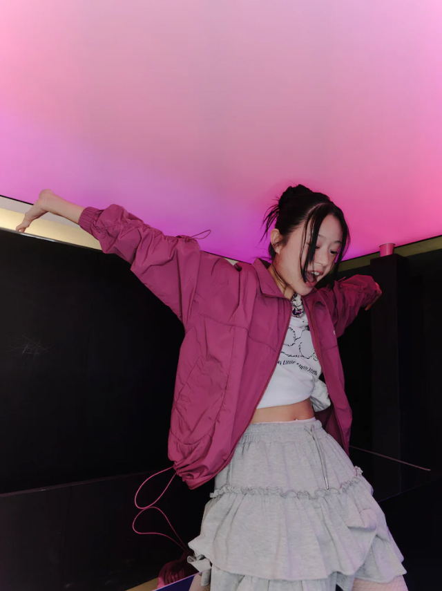 Jisoo Seo poses playfully under a pink-lit ceiling, wearing a magenta windbreaker, cropped graphic tee, and ruffled grey skirt.