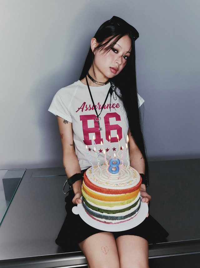 Jisoo sits holding a colorful rainbow-layered birthday cake with lit candles wearing a white graphic tee and a black skirt.