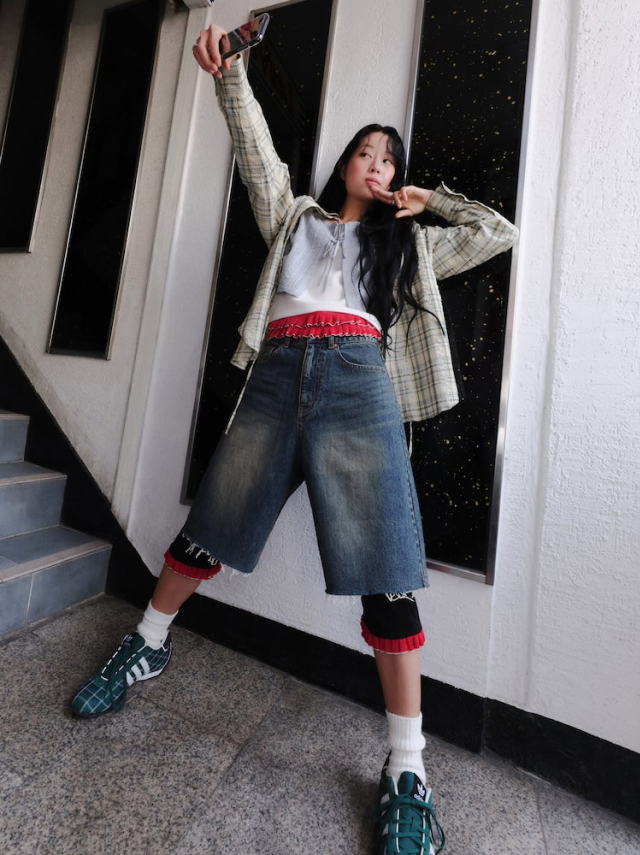 Jisoo Seo poses for an iPhone selfie, wearing an oversized plaid flannel, a cropped grey knit, jorts, green sneakers and layered socks.
