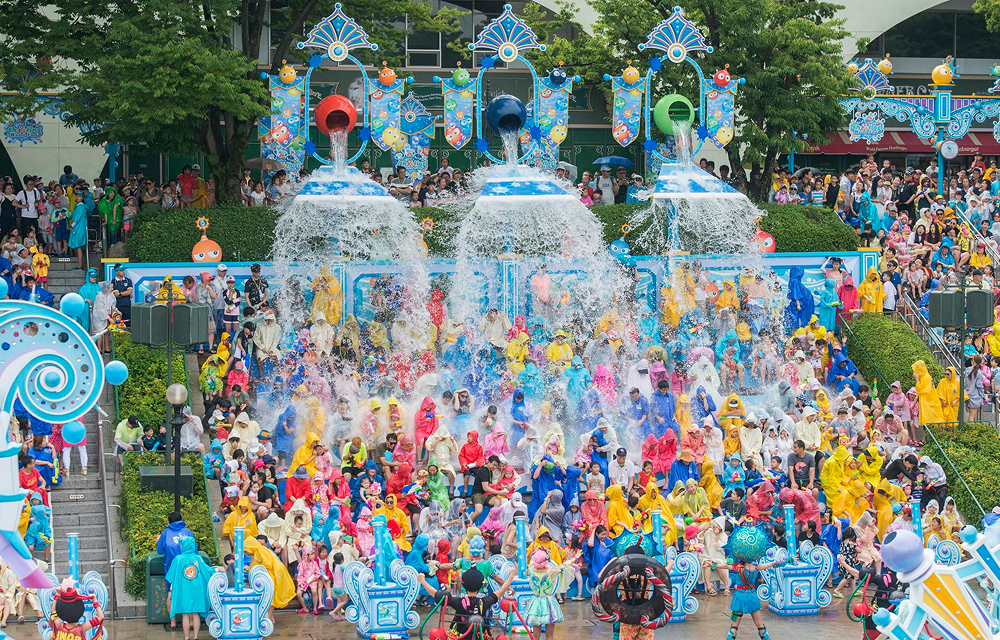 A packed audience dressed in raincoats sits below giant pouring buckets as water splashes down from above. Performers in themed outfits interact with the crowd, while blue and white decor adds to the festive atmosphere.