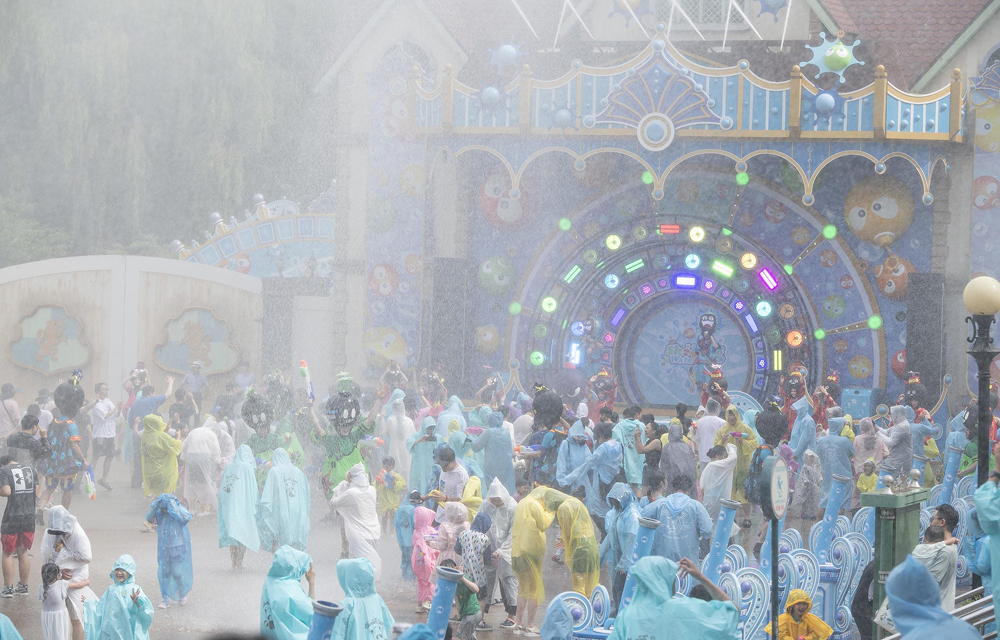 Families and children in colorful rain ponchos play in front of a decorative stage with bright LED lights and cartoon designs. Water sprays from above as the crowd gets soaked during the performance.