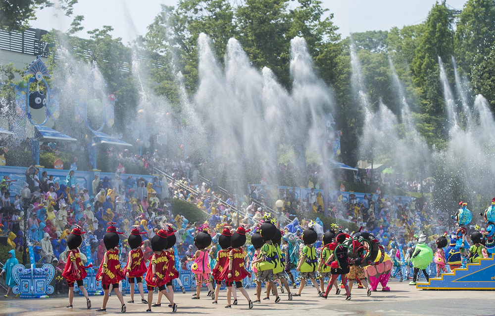 A colorful outdoor performance at Everland featuring dancers in vibrant costumes and oversized character heads. Behind them, fountains shoot water high into the air as crowds in ponchos watch from the stands.