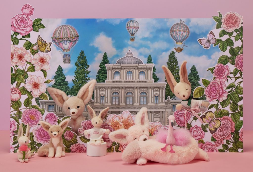 A collection of plush desert fox, bunny, and flamingo toys arranged in front of a rose-themed illustrated backdrop featuring a castle and hot air balloons.
