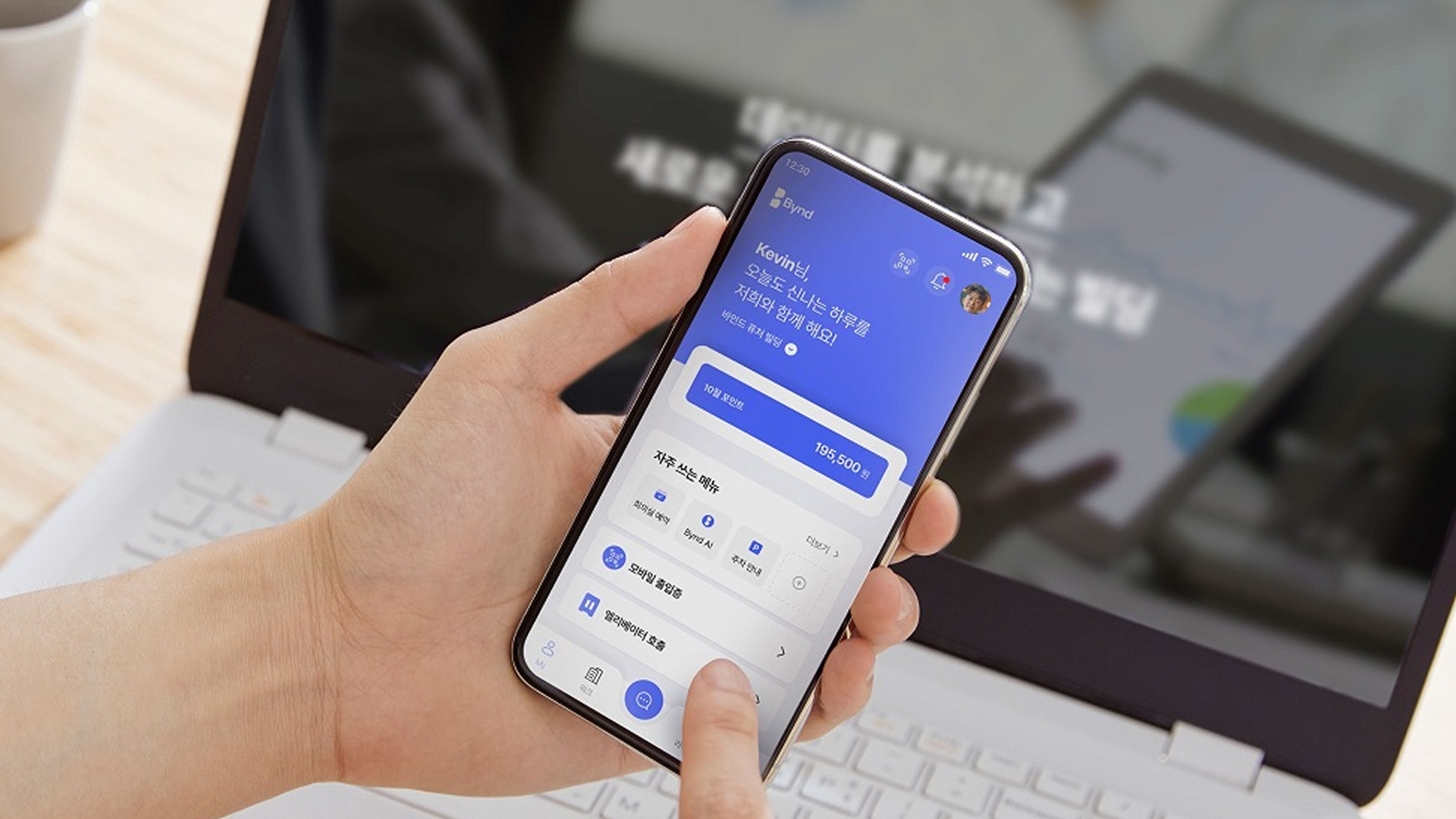 A person holding a smartphone displaying the Bynd app interface in front of a laptop, illustrating Samsung C&T E&C Group’s smart building platform.