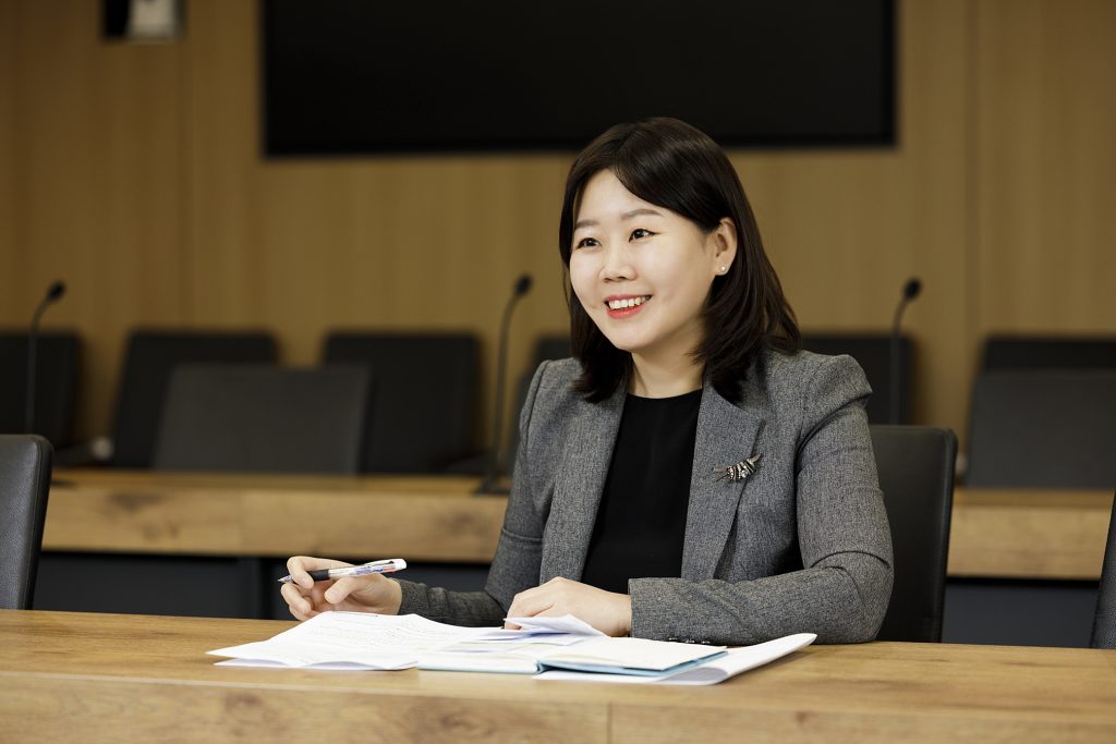 Jungwan Kim sits at a conference table with documents and a pen, smiling during a meeting in a modern boardroom.