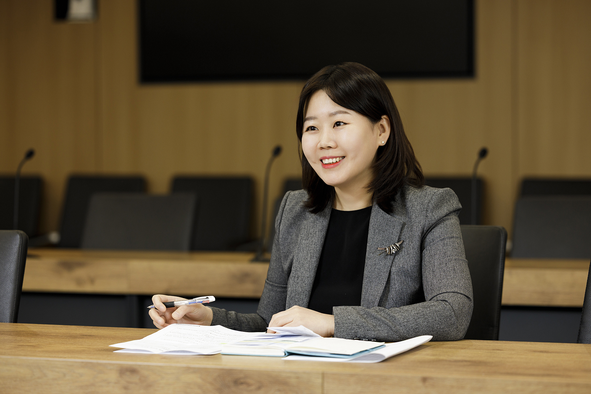 Jungwan Kim sits at a conference table with documents and a pen, smiling during a meeting in a modern boardroom.