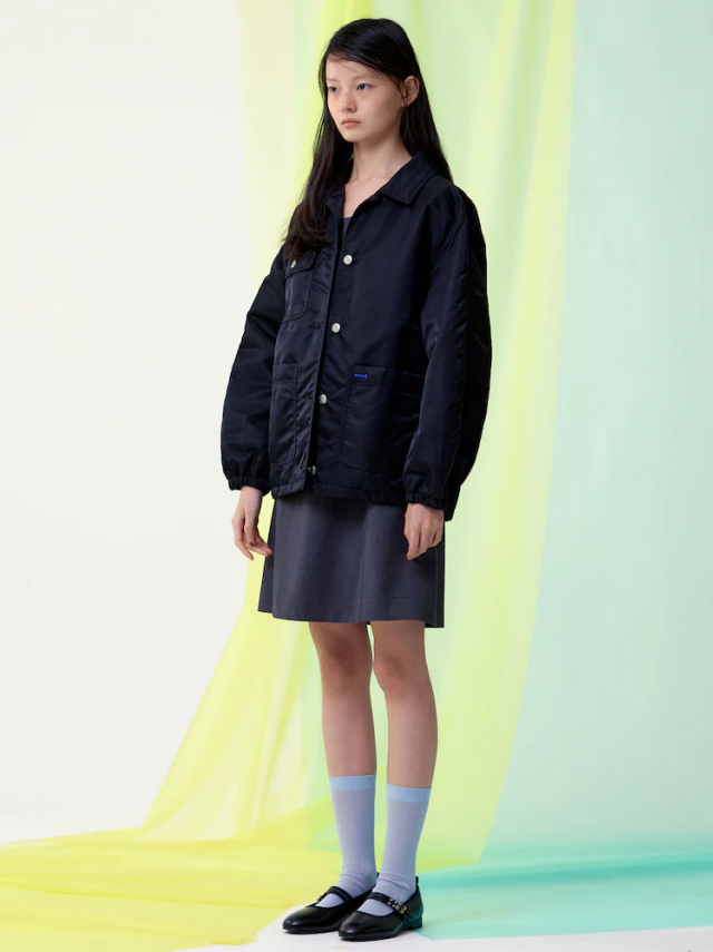A model wears a navy jacket and grey skirt against a pastel yellow fabric backdrop, wearing black KUHO mary jane shoes with light blue socks.