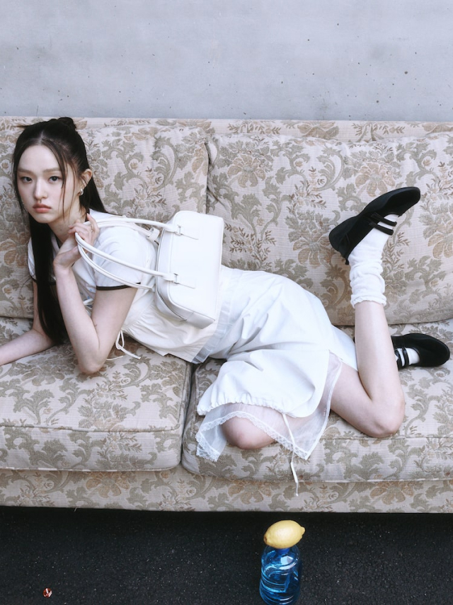 A woman on a vintage floral sofa wears a layered white skirt and a white blouse with black Mary Jane sneakers and a double velcro strap. The look is accessorized with white socks and a small white bowling bag.