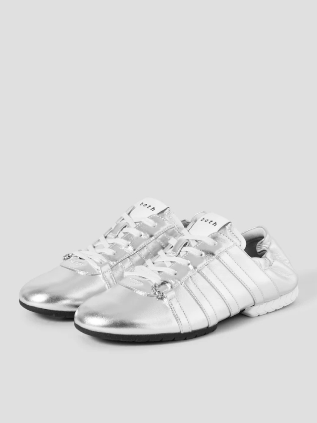 Sneakerinas from the brand BOTH; silver sneaker-style shoes with shiny finish, white laces, side stitching, and a heel that resembles the chevron patterns seen on a car tyre.