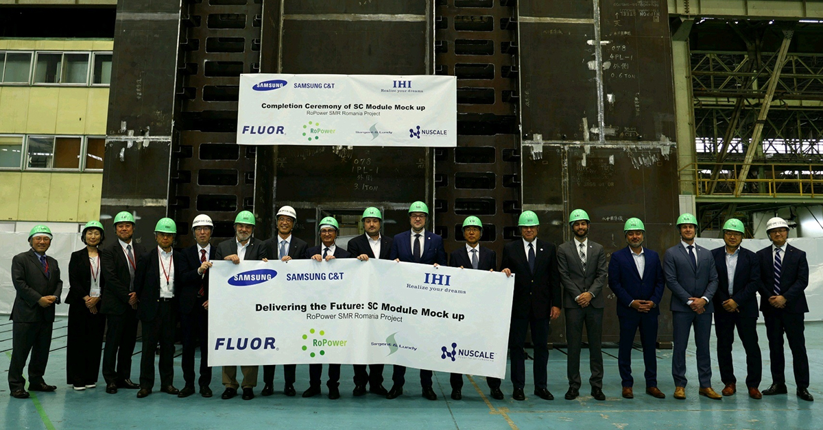 A group of representatives from the Romanian SMR Project pose for a photo in front of a large SC module at IHI's Yokohama plant. They hold banners reading "Completion Ceremony of SC Module Mockup" and "Delivering the Future: SC Module Mockup" for the RoPower SMR Romania Project.