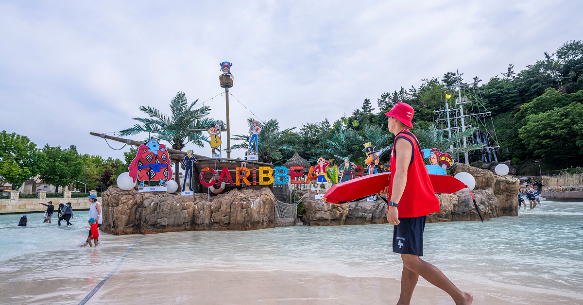 Lifeguard walking through shallow water in front of a colorful Caribbean Bay sign decorated with One Piece characters.