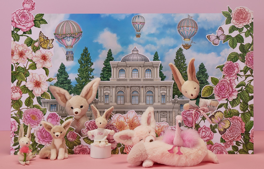 A collection of plush desert fox, bunny, and flamingo toys arranged in front of a rose-themed illustrated backdrop featuring a castle and hot air balloons.