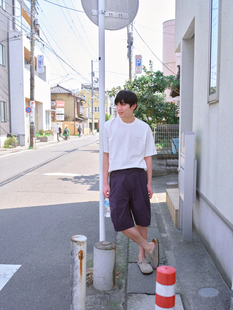 Model wears navy chino shorts with a loose white tee, standing against a pole-lined sidewalk on a bright summer day.