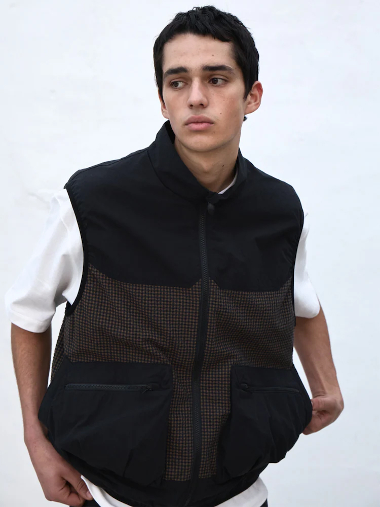 Model wears a black and brown utility vest with oversized cargo trousers and brown derbies, standing against a pale wall.