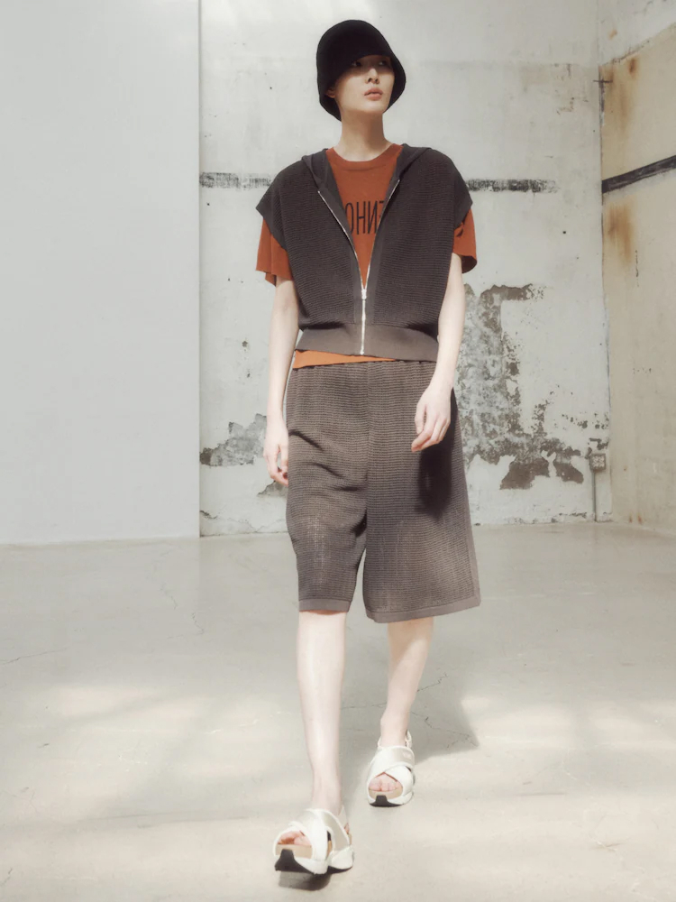 Model wears KUHO's wide knit shorts in ash gray with a matching vest and oversized tee, walking in a sunlit industrial space.
