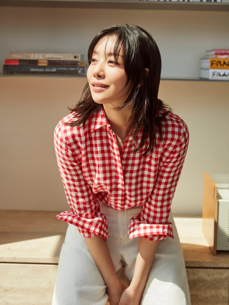 Model wears the Beanpole red striped short-sleeve shirt with white pleated shorts, seated beside a potted plant in bright daylight.