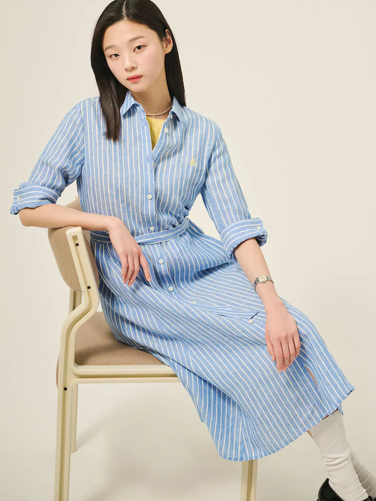Female model wears the Beanpole blue linen stripe shirt dress with rolled-up sleeves, seated on a cream chair in a minimal studio space.
