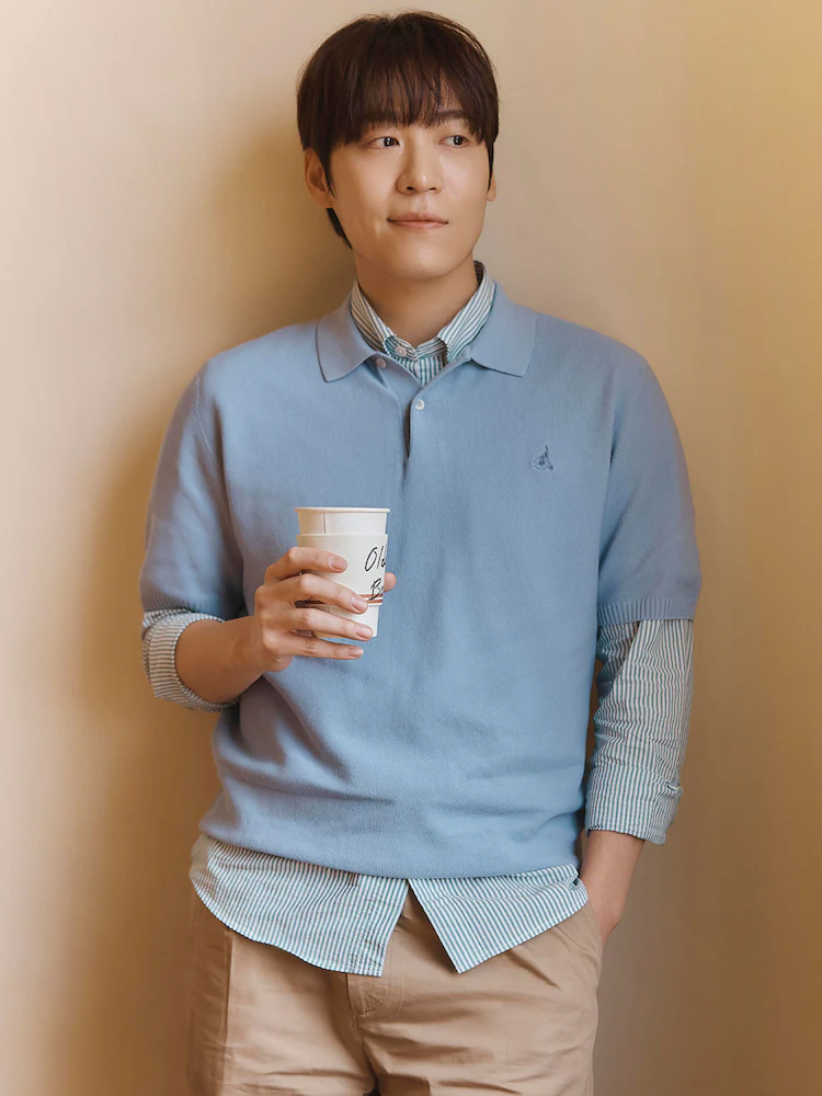 Male model wears the Beanpole sky blue cotton-silk knit over a green striped shirt, styled with beige trousers and a takeaway coffee cup.