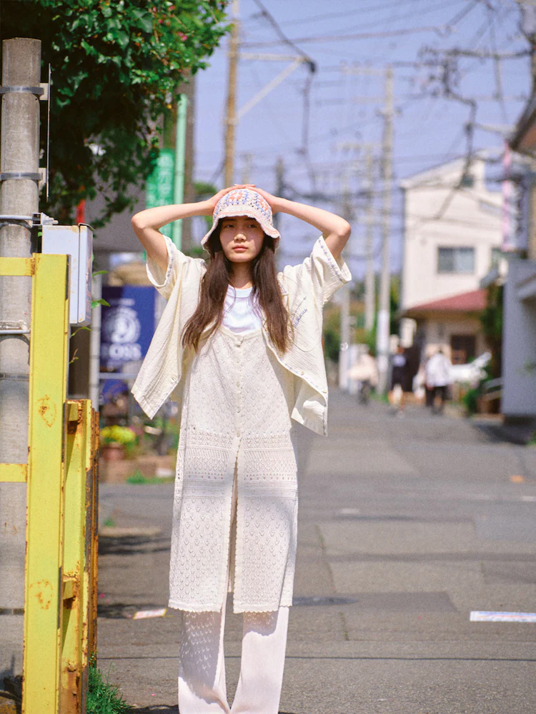 Model wears the Beaker crochet knit dress layered over wide-leg pants, paired with a crochet top and bucket hat, standing on a quiet street.