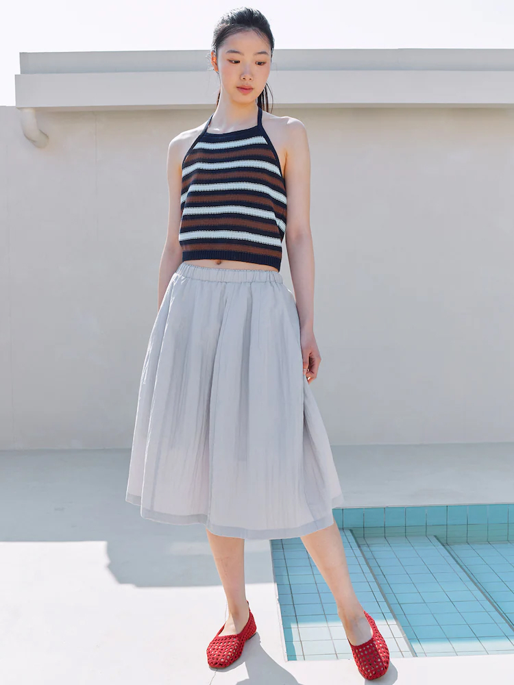 Model wears the KUHO Plus Sheer Volume Skirt in light grey with a striped halter knit top and red woven flats by a pool.
