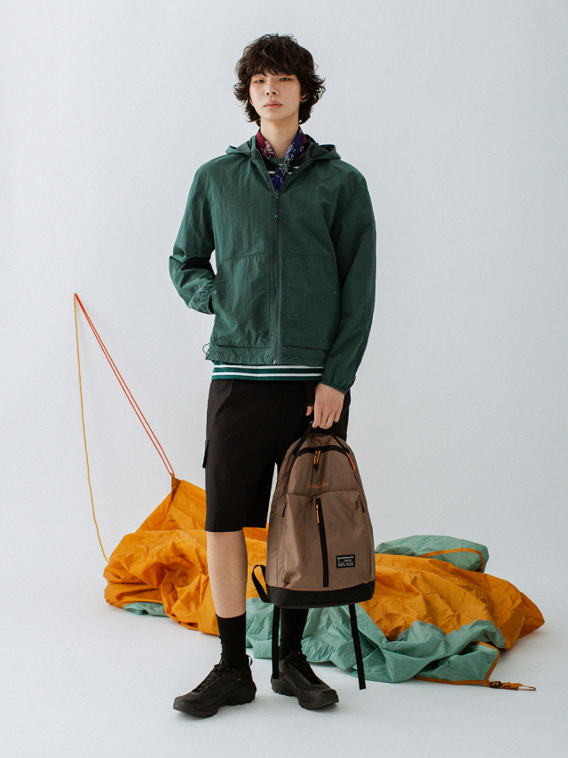 A man poses indoors in front of a white backdrop and colorful parachute fabric, wearing a green windbreaker, black shorts, black socks, and black sneakers. He carries a brown backpack.