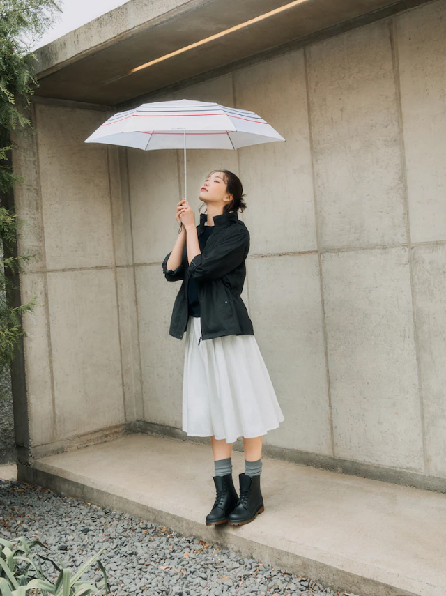 A woman stands in front of a concrete wall, gazing upward while holding a white umbrella with red stripes. She wears a black rain jacket, a white skirt, gray socks, and black lace-up rain boots.
