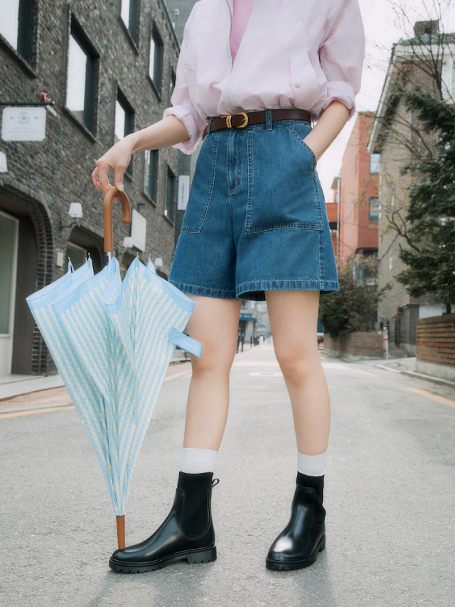 A woman in a pink blouse and denim shorts stands on a city street holding a blue gingham umbrella. She wears black rain boots and white socks.