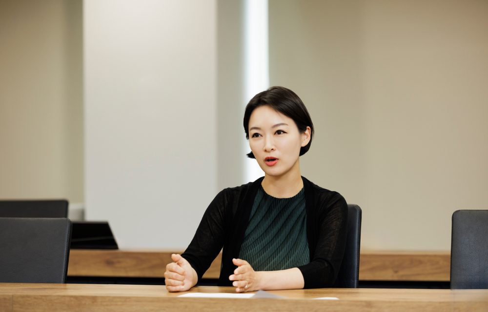Soh-young Cecilia Lim seated at a conference table, actively speaking and gesturing with both hands during an interview.