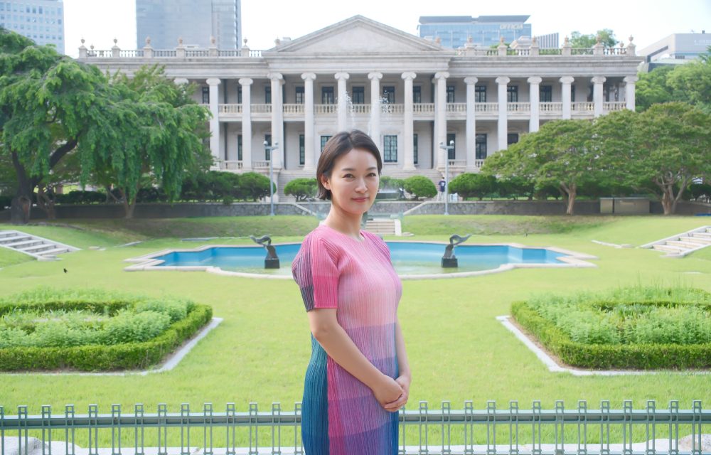 Soh-young Cecilia Lim stands in a garden in front of a historic building, wearing a pink and blue dress and smiling at the camera.