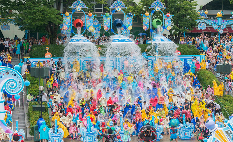 Large crowd in colorful raincoats getting drenched by overhead splash buckets during a water performance.