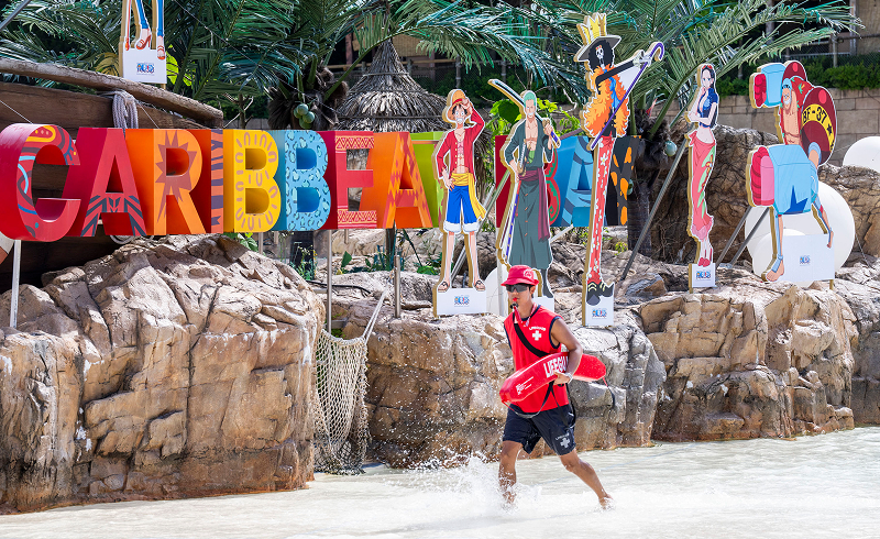 Lifeguard in red uniform running through shallow water with One Piece character cutouts and Caribbean Bay sign in the background.
