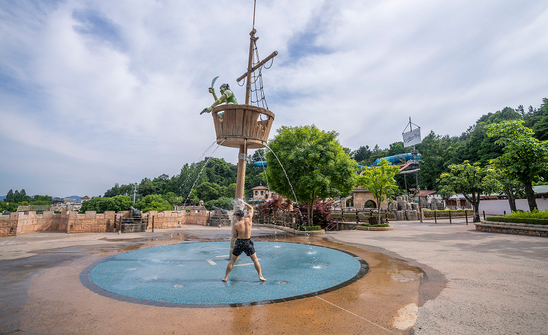 Man cooling off under water sprays from a pirate ship-themed fountain at a splash pad.