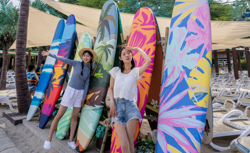 Two young women posing in front of colorful decorative surfboards at a sandy photo zone.