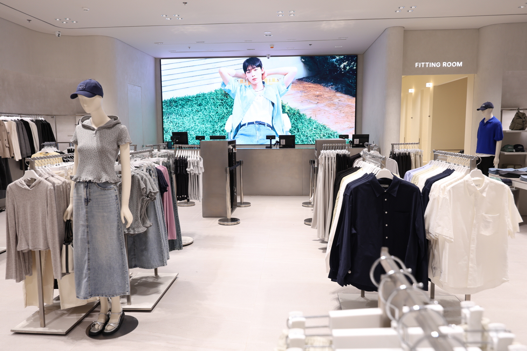 Inside the 8 Seconds store, a large screen behind the checkout counter displays a campaign image of Sunoo reclining on the grass. Clothing racks in the foreground feature casual and neutral-colored garments.