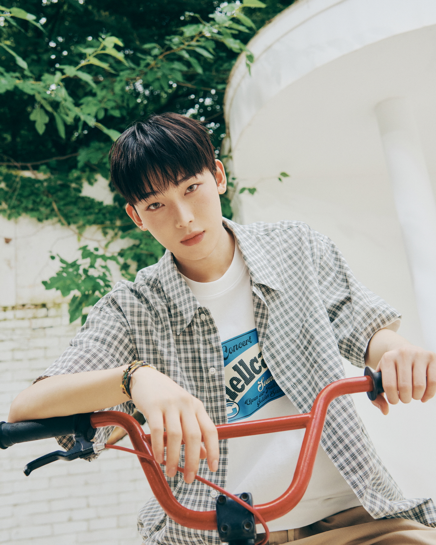 Sunoo leans forward on the handlebars of a red bicycle, wearing a checkered short-sleeve shirt over a white concert-print T-shirt. With greenery in the background, the photo evokes a retro, summery atmosphere.